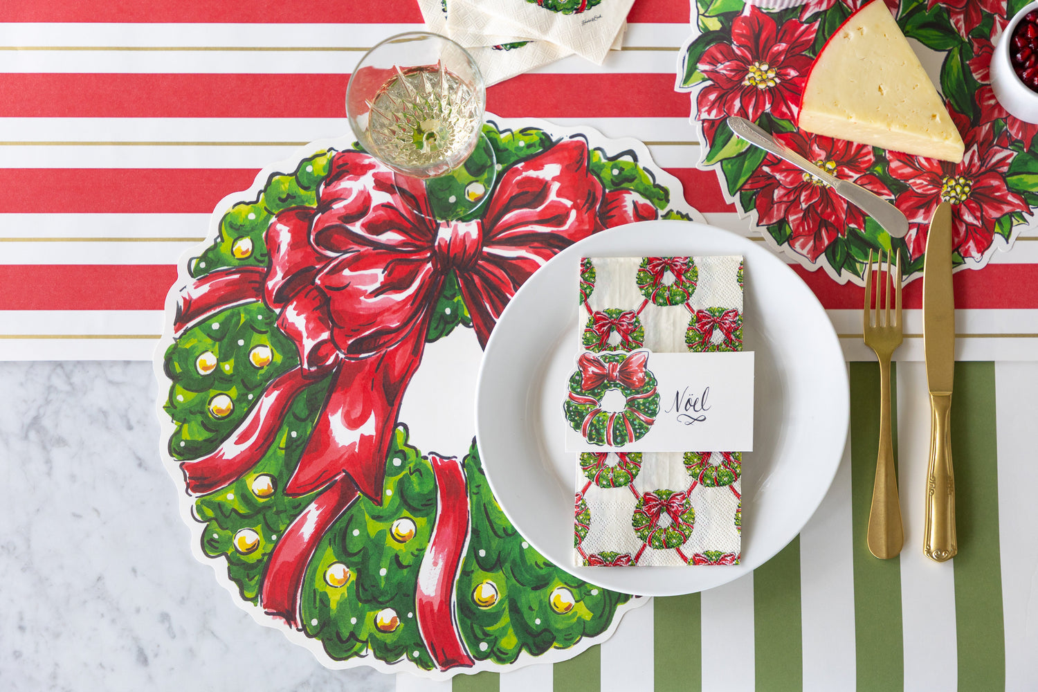 A place setting featuring the Die-cut Holiday Wreath Placemat, a plate with a Holiday Wreath Guest Napkin and Holiday Wreath Place Card atop, Holiday Wreath Serving Paper with a block of cheese, and Red &amp; Gold Awning Stripe Runner underneath.