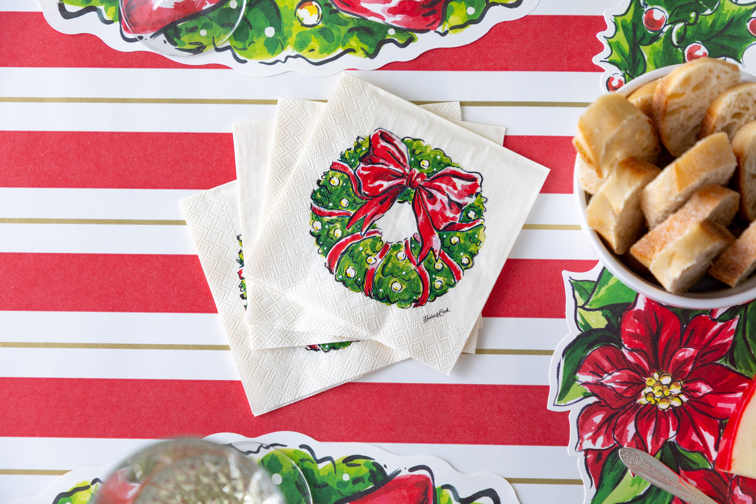 A stack of Holiday Wreath Cocktail Napkins on a festive table setting with the Red &amp; Gold Awning Stripe Runner underneath.
