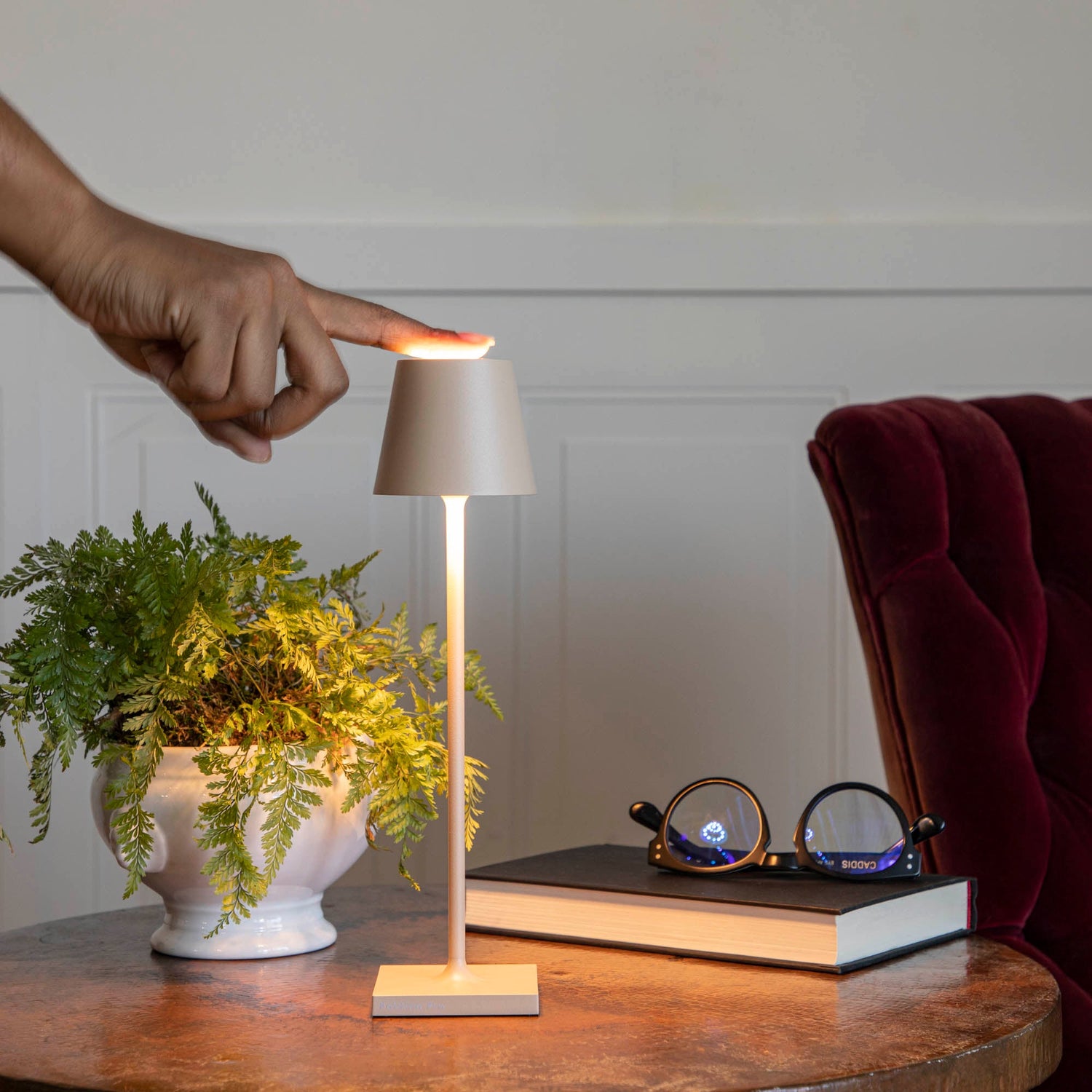 A Zafferano lamp on a side table with a finger touching the top to turn the lamp on. 
