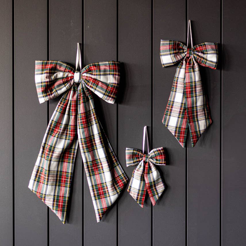 Three plaid fabric bows in red, green, black, and white tartan patterns, displayed on a dark gray vertical wood plank wall.
