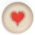 A round beige enamel trinket tray with artwork of a red heart on a white background. 