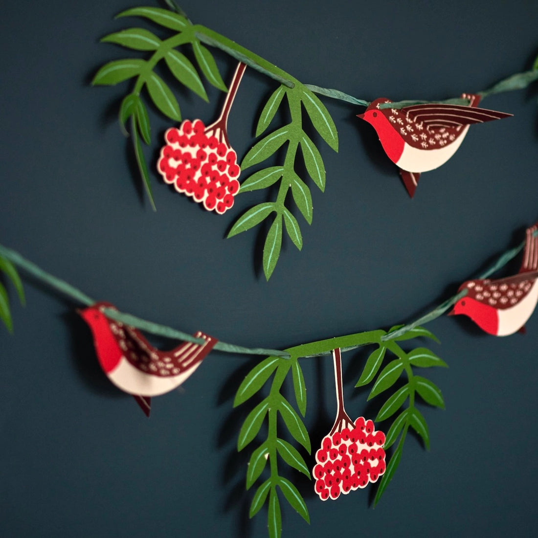 A cut paper garland of 3D birds in red and brown, green leaves, and red berries on a dark teal wall. 