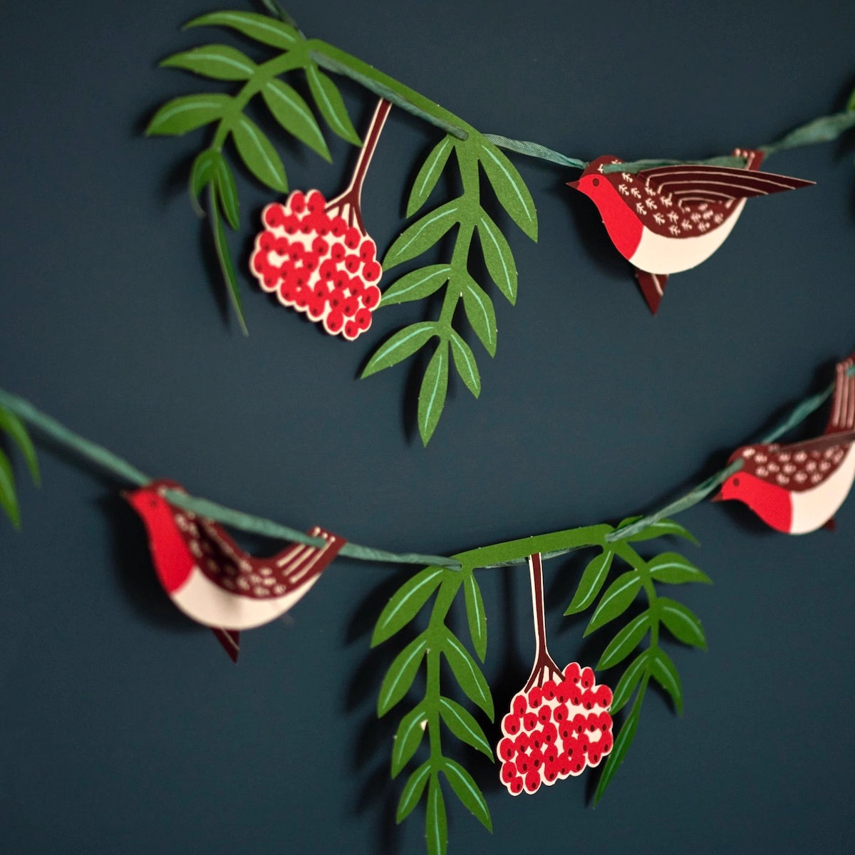 A cut paper garland of 3D birds in red and brown, green leaves, and red berries on a dark teal wall. 