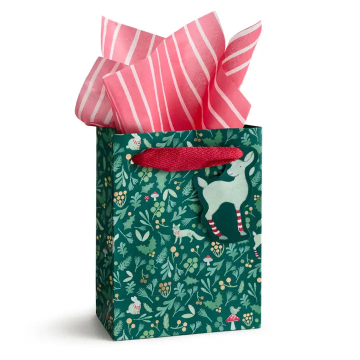 A green gift bag with a festive woodland pattern of deer, leaves, and berries, filled with pink and white striped tissue paper and topped with a red ribbon handle.