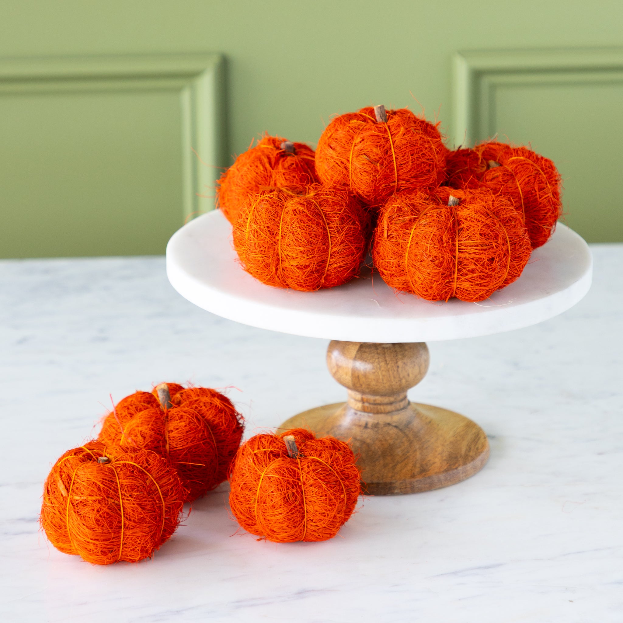 Burnt orange coco fiber pumpkins on a white cake stand with a wooden base against a green wall.