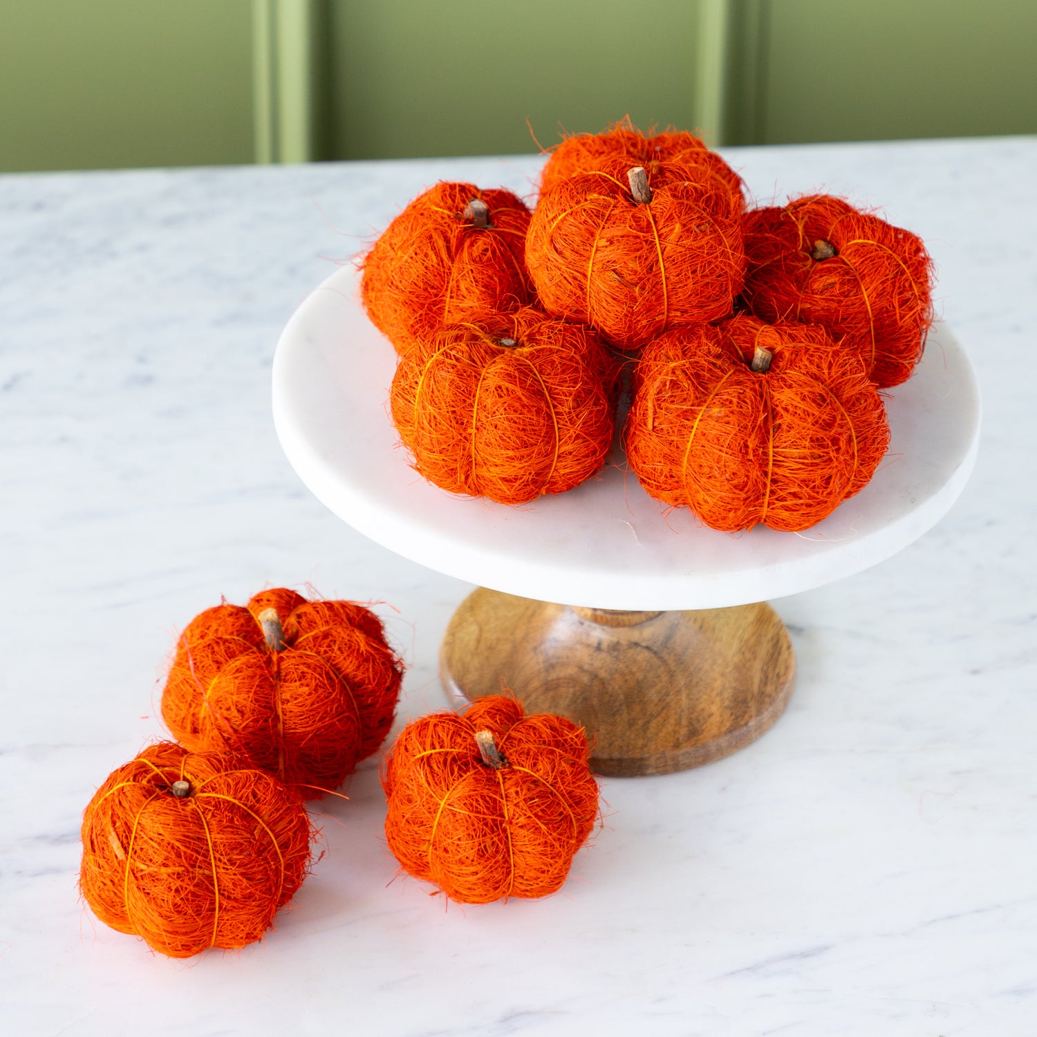 Burnt orange coco fiber pumpkins on a white cake stand with a wooden base against a green wall.