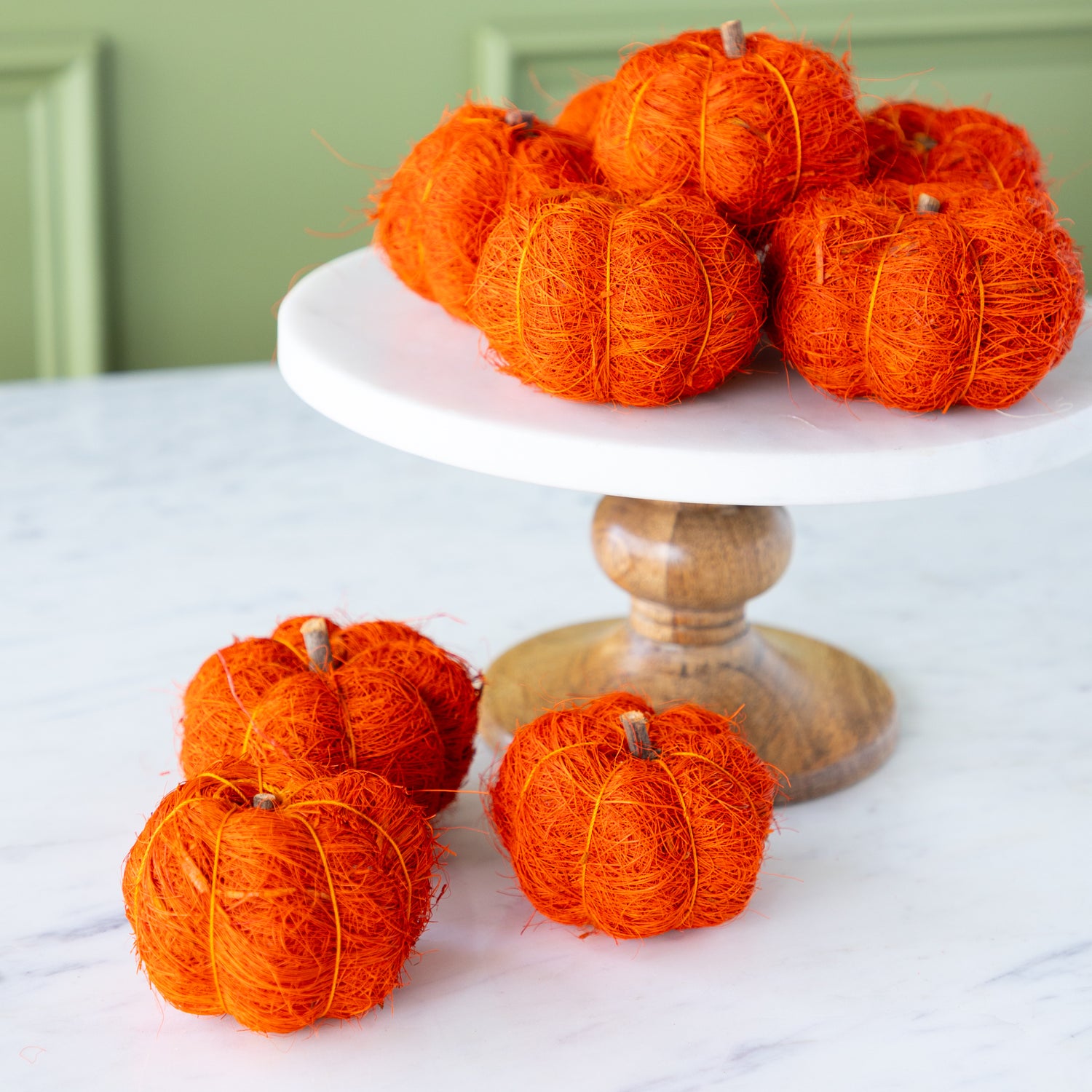 Burnt orange coco fiber pumpkins on a white cake stand with a wooden base against a green wall.