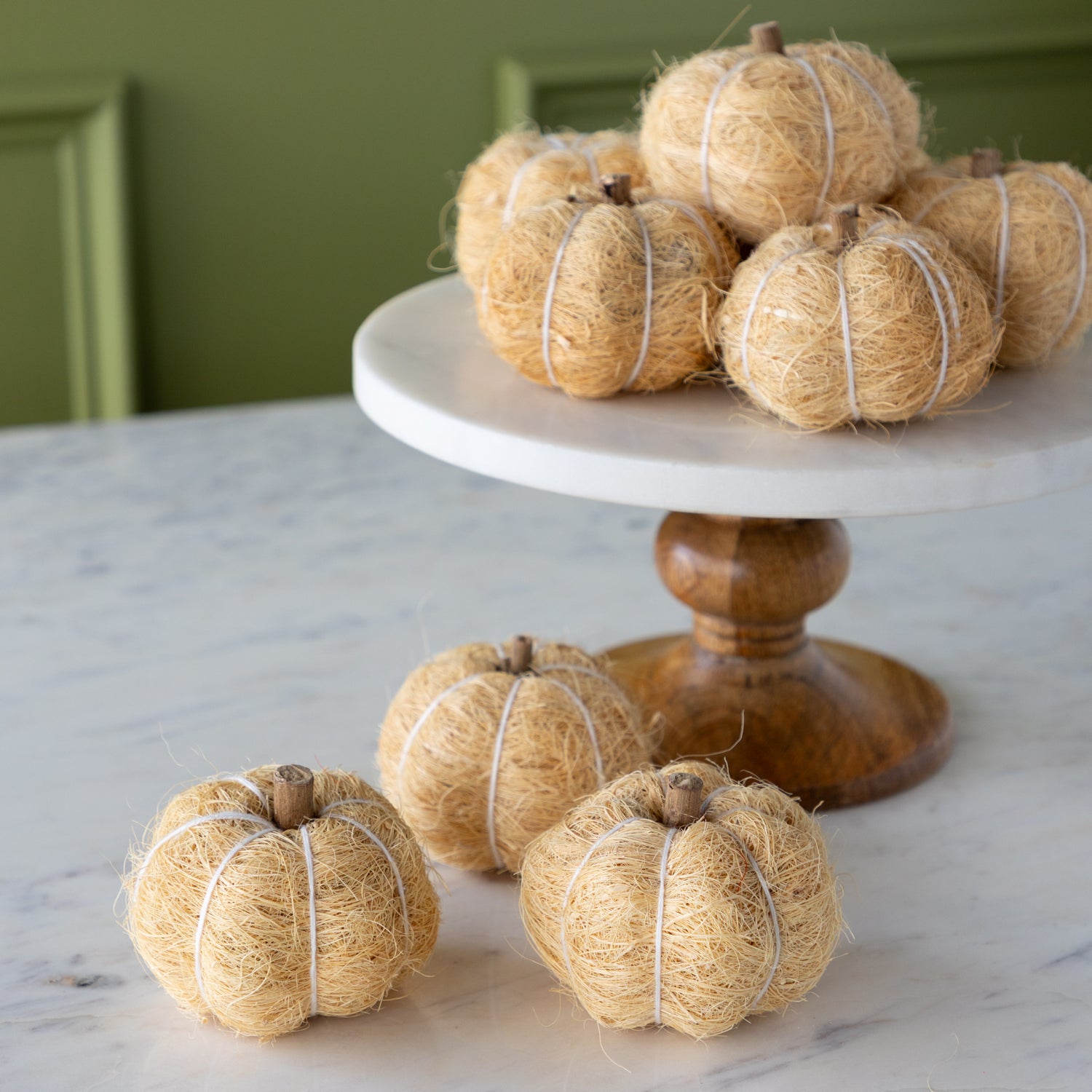 Cream coco fiber pumpkins on a white cake stand with a wooden base against a green wall.