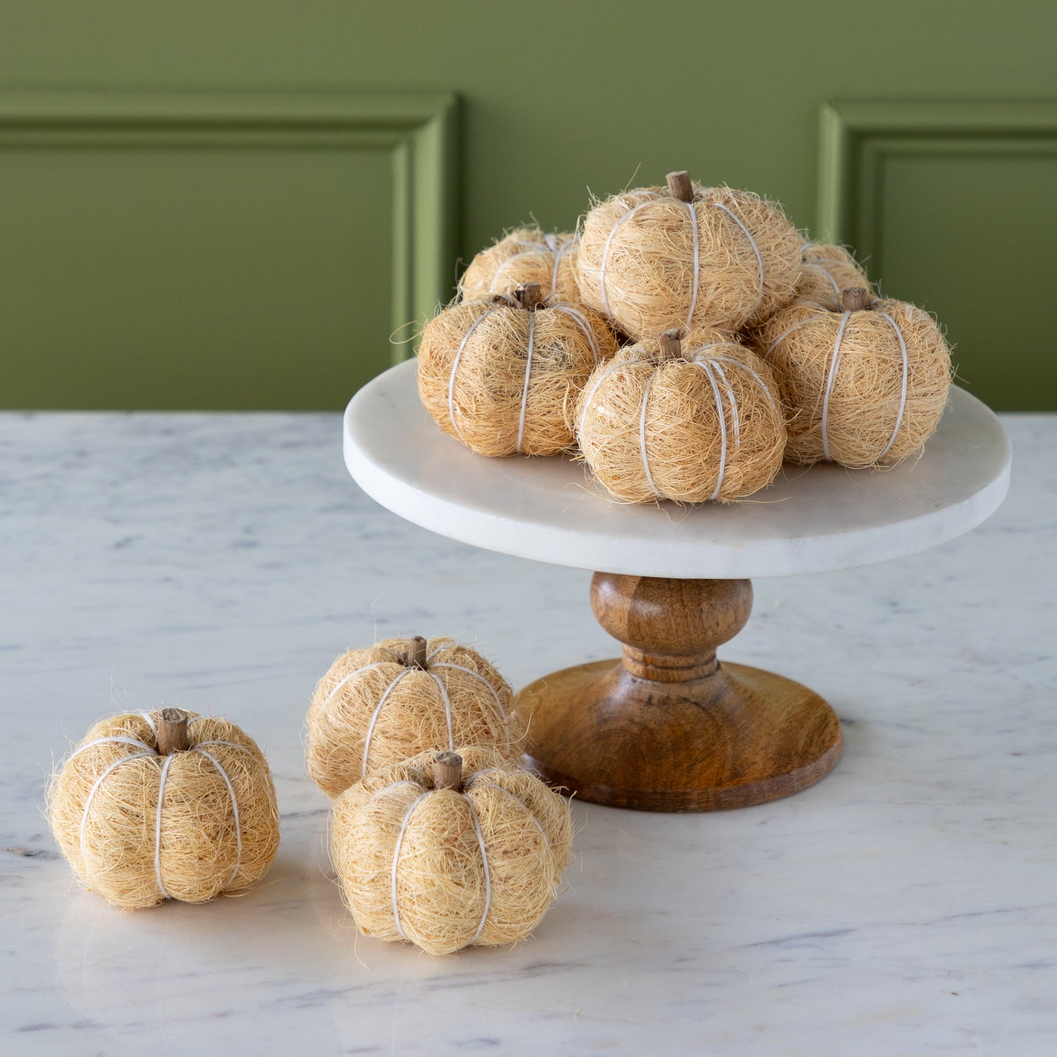Cream coco fiber pumpkins on a white cake stand with a wooden base against a green wall.