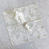 A square white napkin with a sage green toile pattern, folded and displayed on a light gray marble surface with a matching napkin ring holding a folded white napkin.