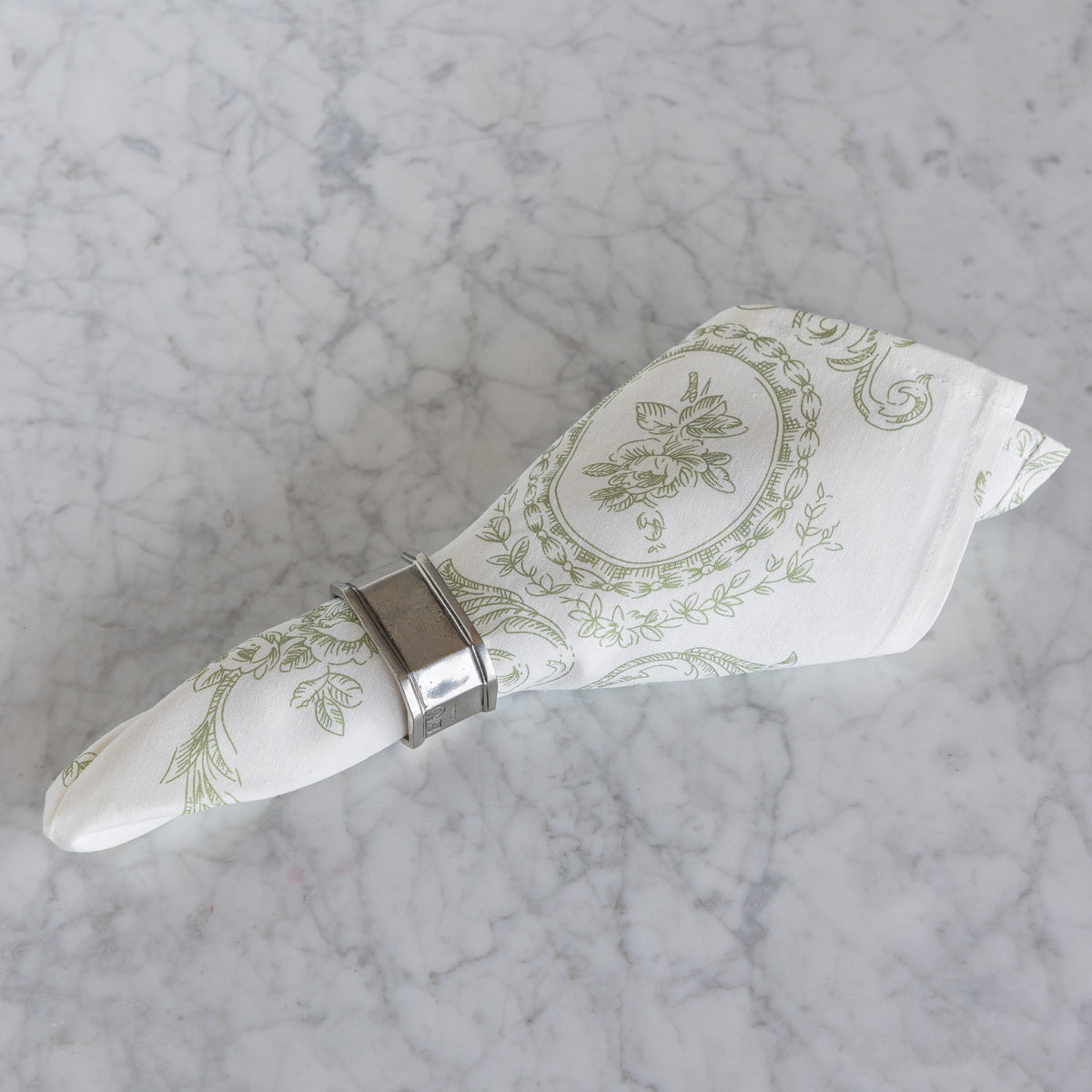 An off-white linen napkin with a sage green botanical pattern, rolled and secured with a silver metal napkin ring, set on a white and gray marble surface.