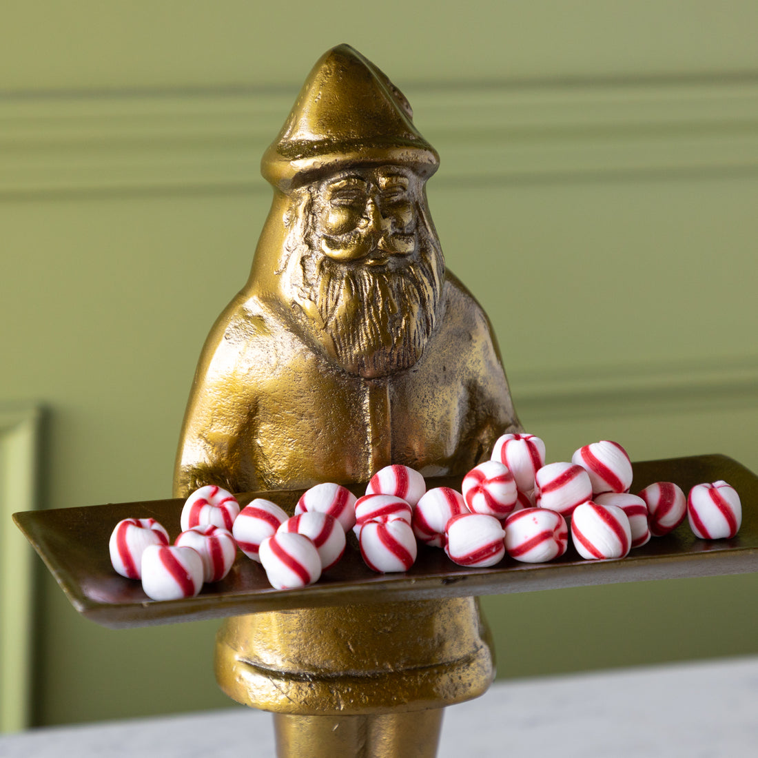 Gold statue of a Saint Nick holding a tray of peppermint candies against a green wall.