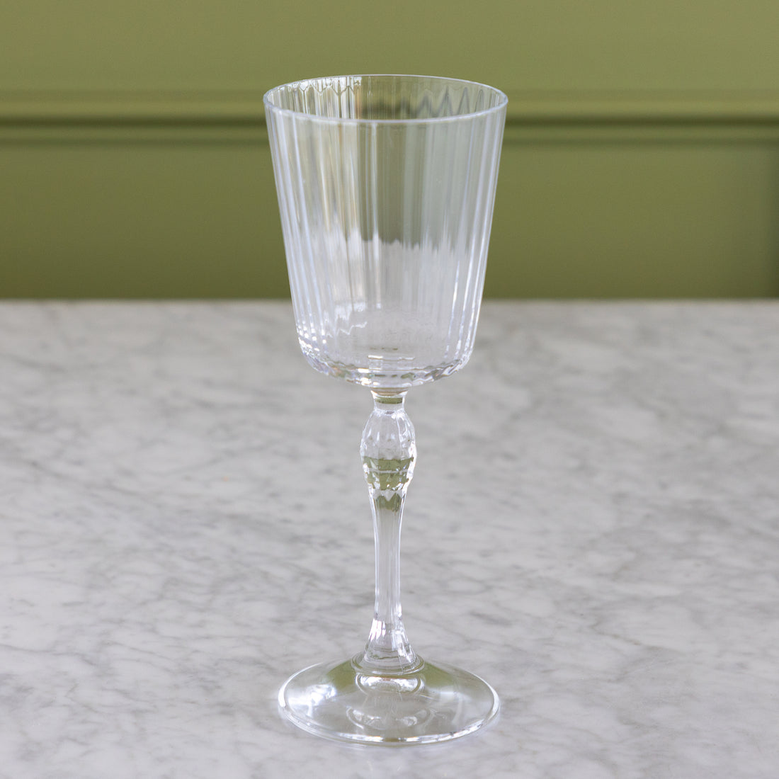 A clear ribbed glass goblet with a flared base, shown standing on a light marble surface against an olive green wall.