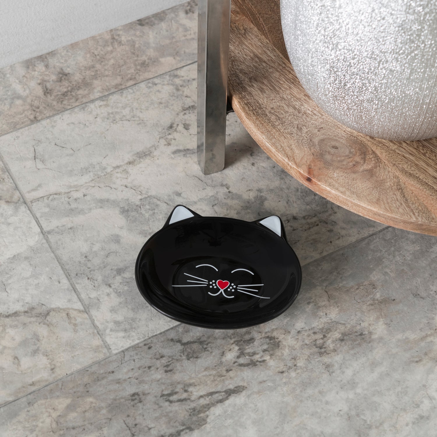 A black ceramic cat-shaped shallow dish with white and red facial details, placed on a gray marble tile floor next to a wooden table leg.