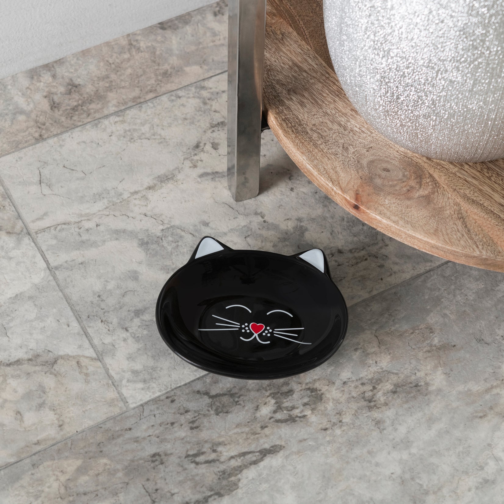 A black ceramic cat-shaped shallow dish with white and red facial details, placed on a gray marble tile floor next to a wooden table leg.