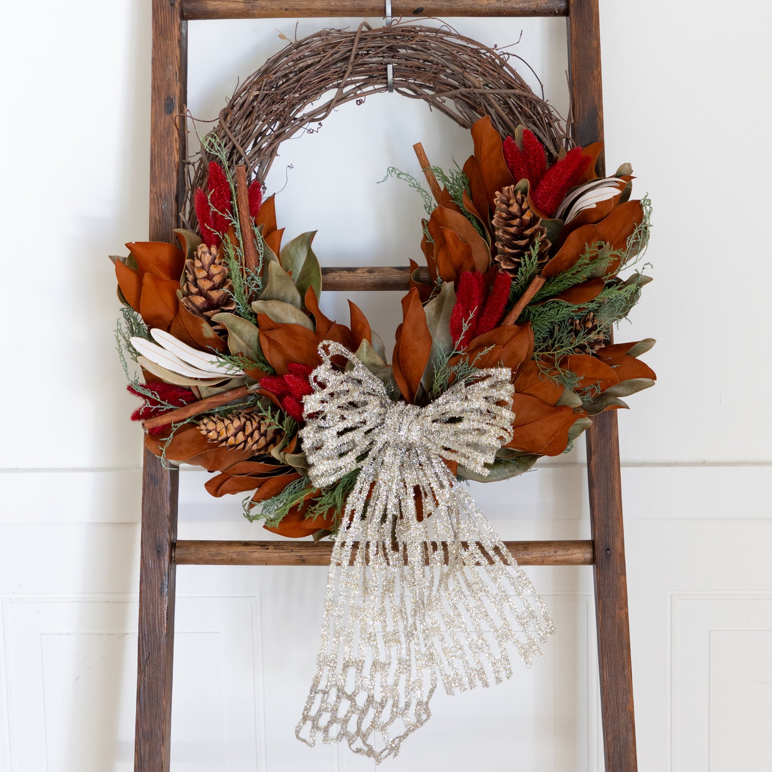 Decorative wreath with autumn leaves and pinecones on a wooden ladder against a white wall.