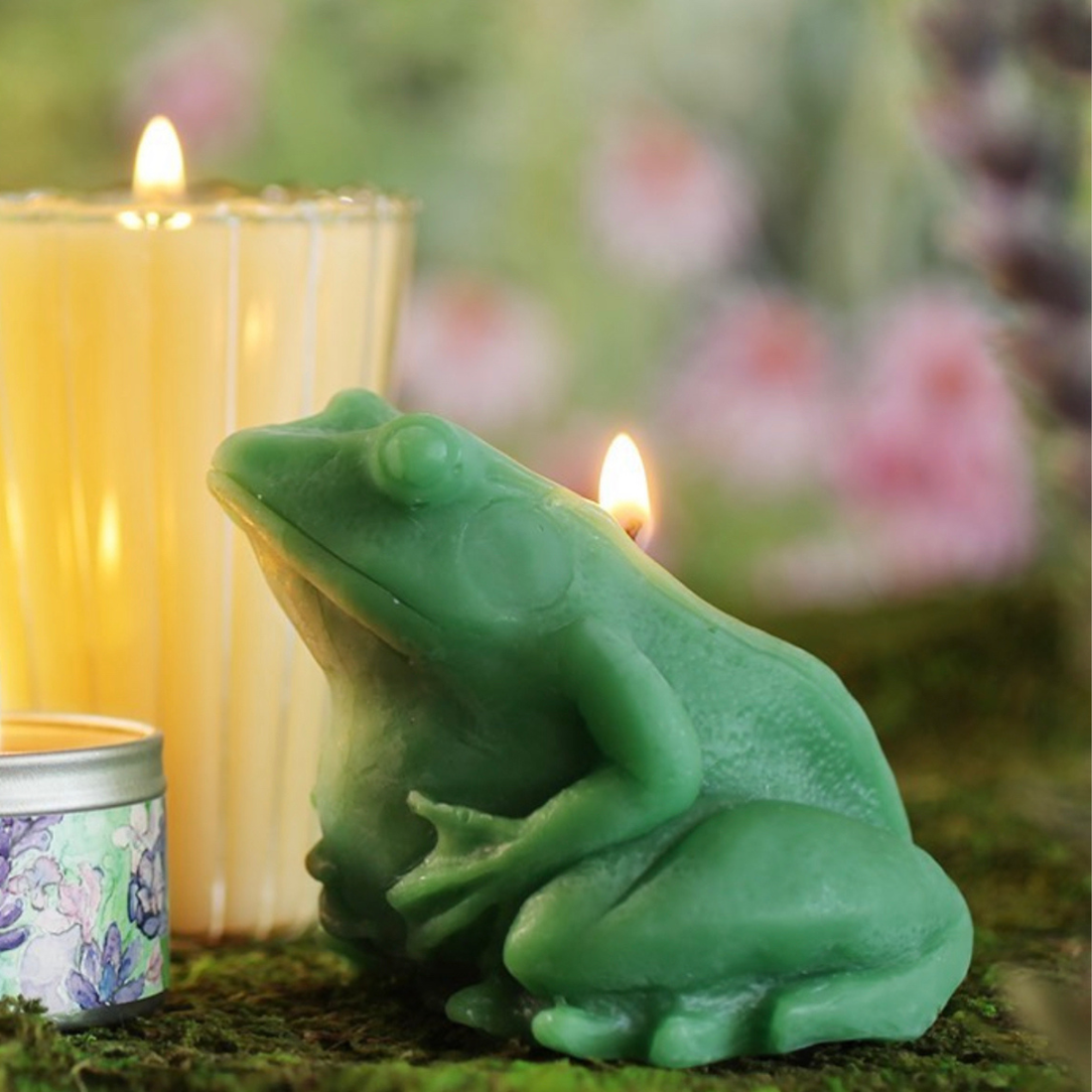 A medium green beeswax candle in the shape of a realistic frog holding its belly. The candle is lit, sitting on a mossy green table with other candles.