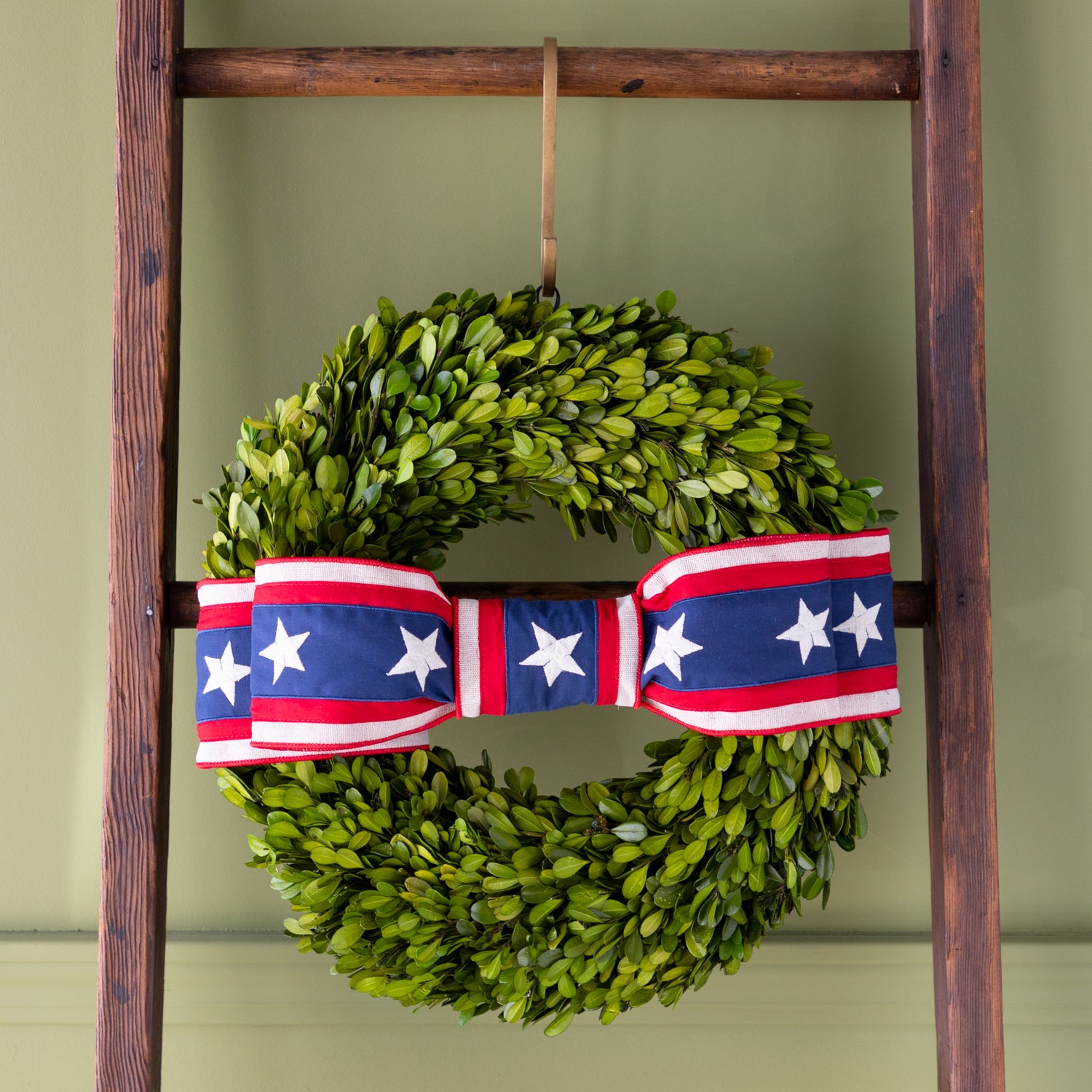 Green wreath with a red, white, and blue ribbon tied in a bow on a wooden ladder against a green wall.