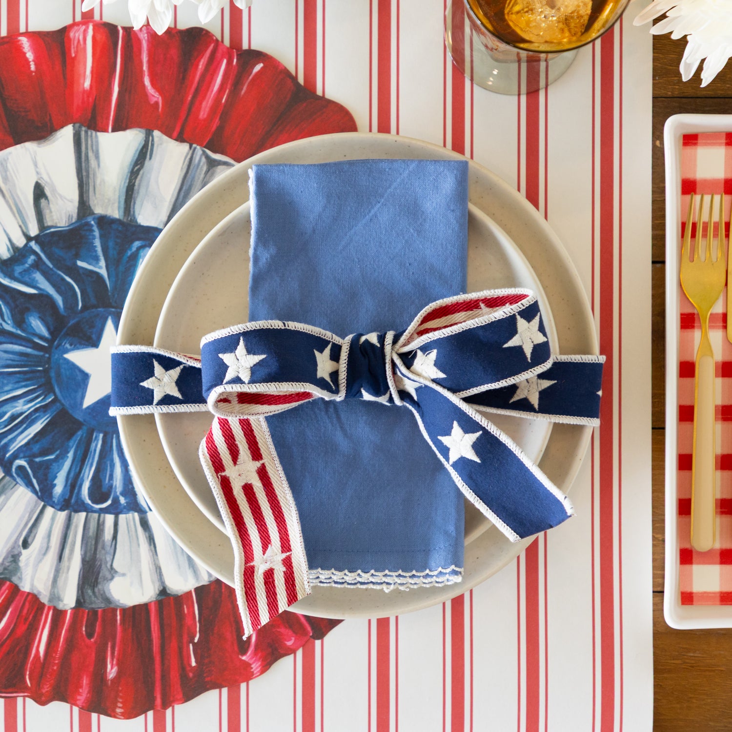 Decorative table setting with red, white, and blue ribbon tied in a bow around a  napkin on a striped table runner.