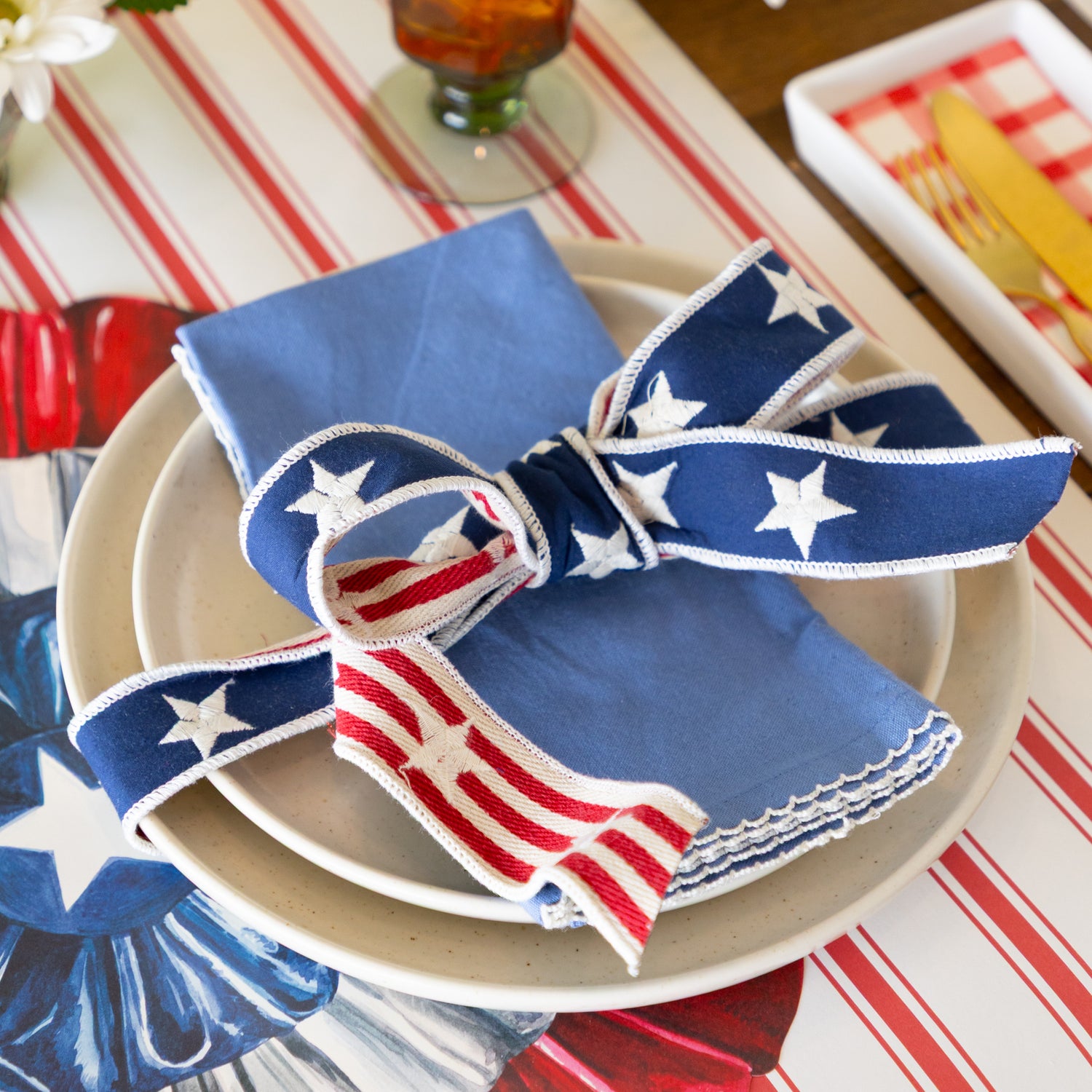 Napkin with American flag patterned ribbon tied around it on a plate with a striped table runner in the background.