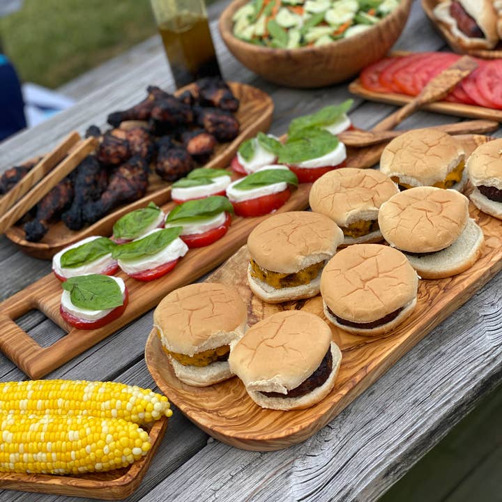A picnic table with various foods on it all being served on olive wood platters and trays. 