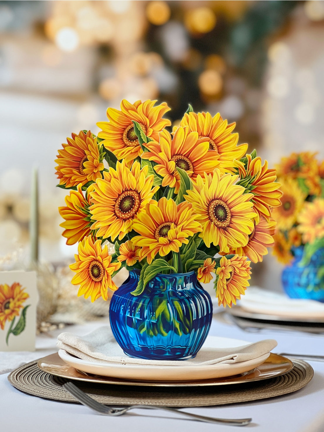 Bouquet of paper sunflowers in a blue vase on a table setting with plates and cutlery.
