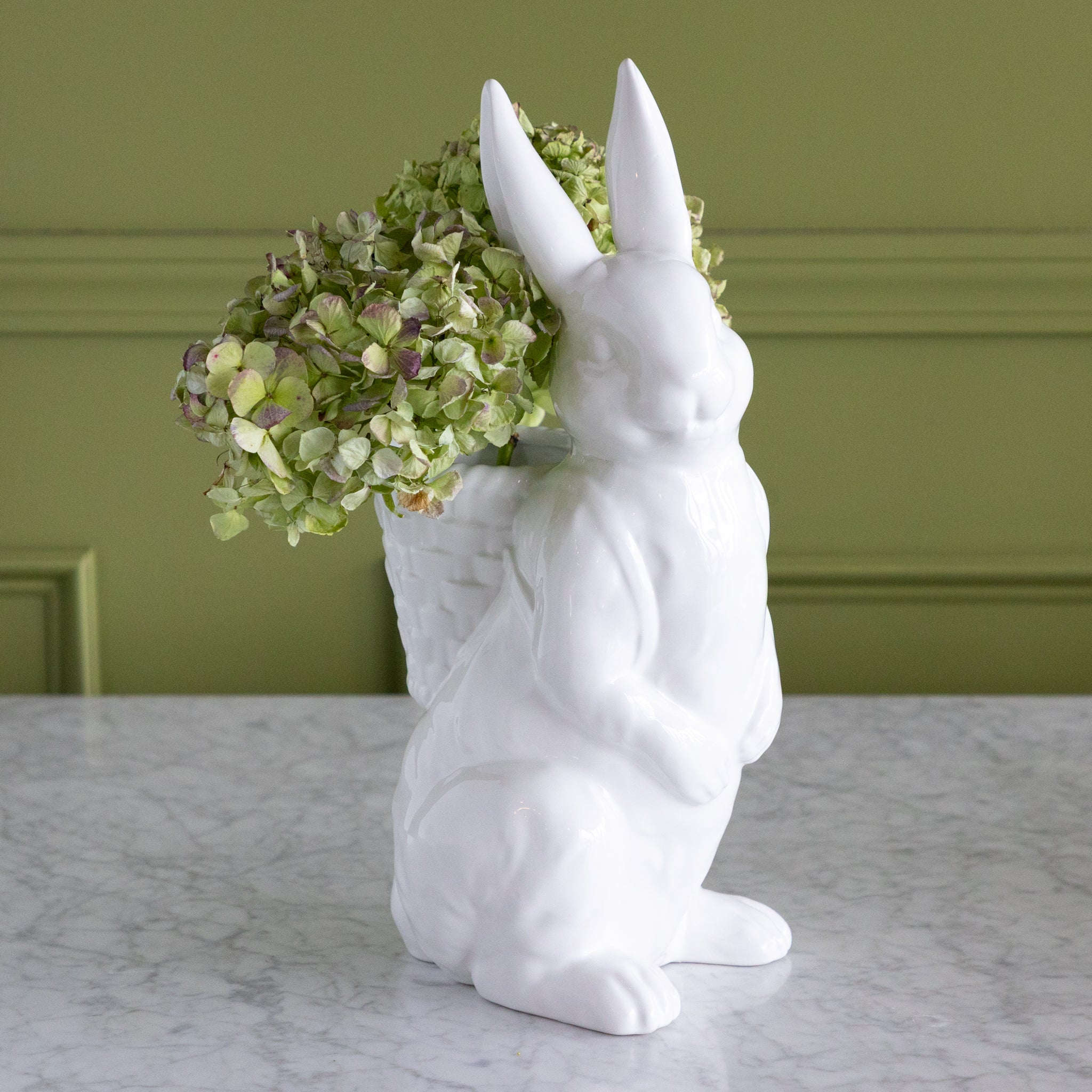 White rabbit-shaped vase with green hydrangeas against a green wall.