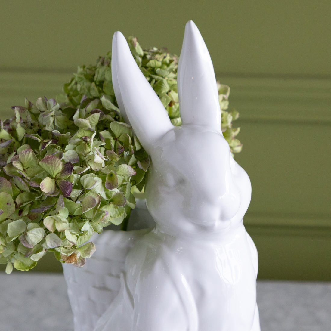 White rabbit-shaped vase with green hydrangeas in its basket against a green wall.