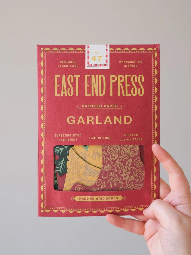 A hand holds a red package of East End Press Garland Printed Paper against a plain light gray background. The package features gold lettering and a small clear window showing folded patterned paper inside.