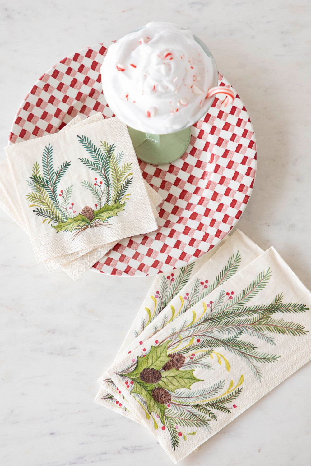 A top-down view of a table with a stack of Christmas Sprigs Cocktail Napkins and a stack of Christmas Sprigs Guest Napkins, fanned out next to a red and white plate and a cup of hot chocolate.