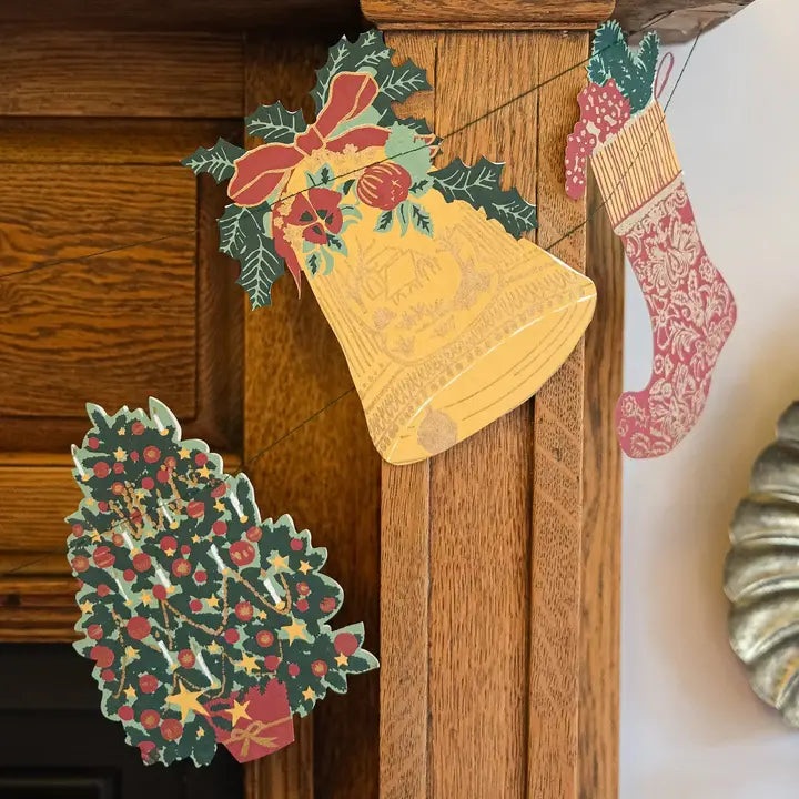 A close-up of a wooden mantel decorated with layered Christmas paper cutouts, including a large gold bell, a red and green bow with holly, a patterned Christmas stocking, and a holly-and-tree motif.
