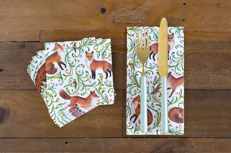 A stack of Fanciful Fox Cocktail napkins and a single Fanciful Fox Guest Napkin on a wooden table with cutlery. 