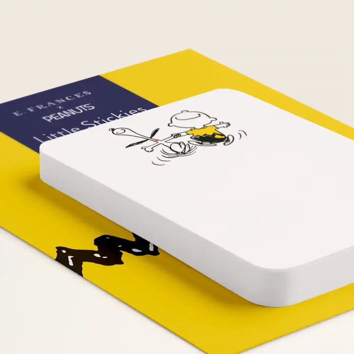 A white rectangular notepad with rounded corners, featuring a black-and-yellow Peanuts cartoon illustration on the cover, resting on a yellow book or folder with a navy title panel that reads &