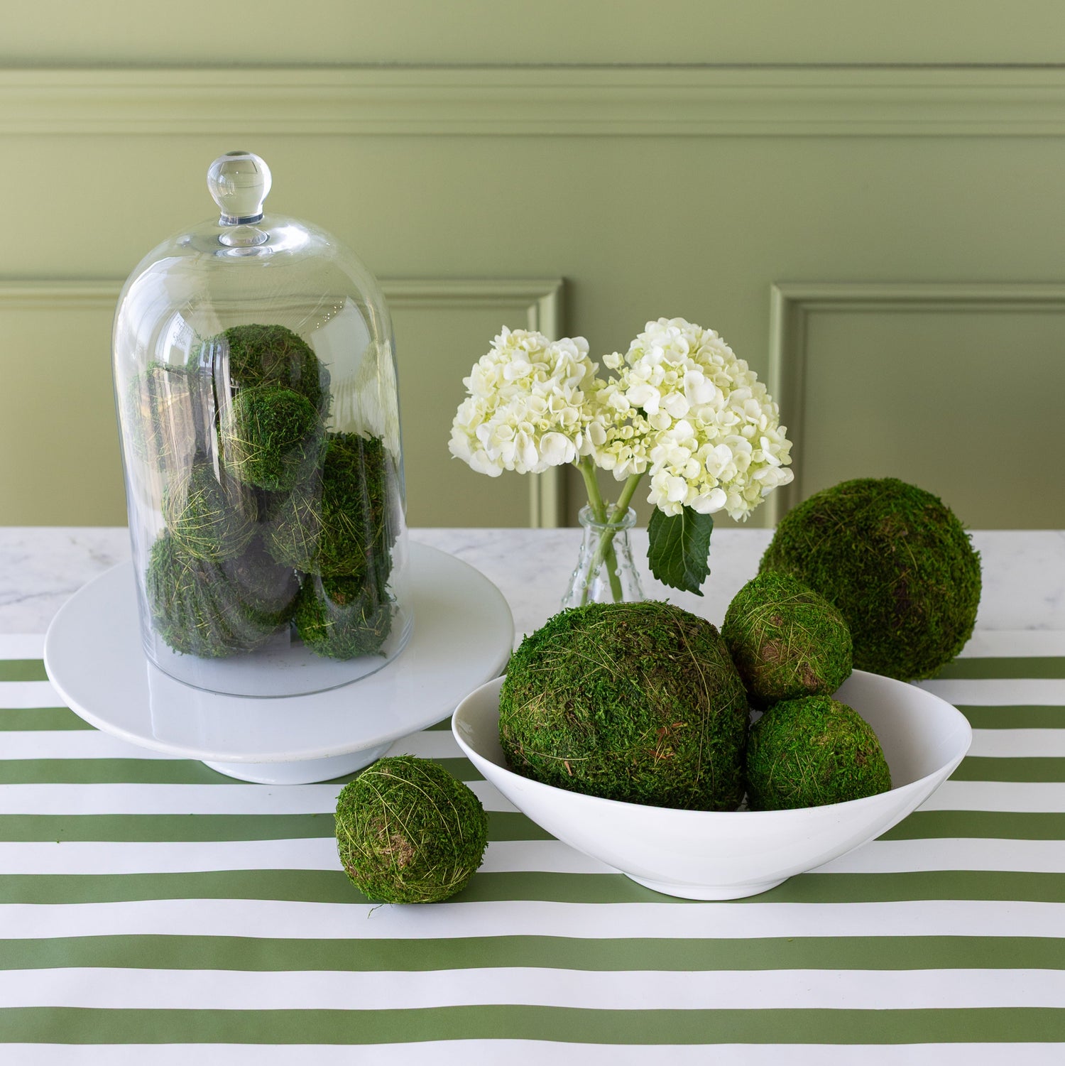 Moss balls displayed in a white bowl on a table with a striped tablecloth, with a glass cloche containing more moss balls in the background, alongside a vase with flowers.