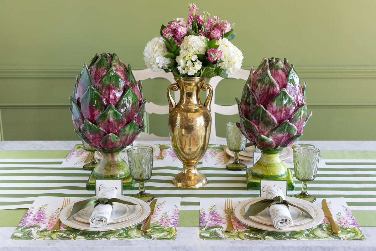 A styled dining table setting featuring two large decorative artichokes on either side of a gold vase filled with pink and white flowers. The table is set with striped green and white table runners, matching floral placemats, white plates, rolled napkins, and clear glassware against a green paneled wall.