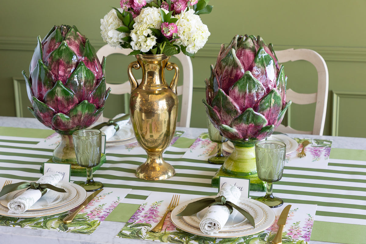 A styled dining table setting featuring two large decorative artichokes in glass vessels, a gold urn vase with white and pink flowers, and matching green-and-white striped table linens with floral placemats and rolled napkins.