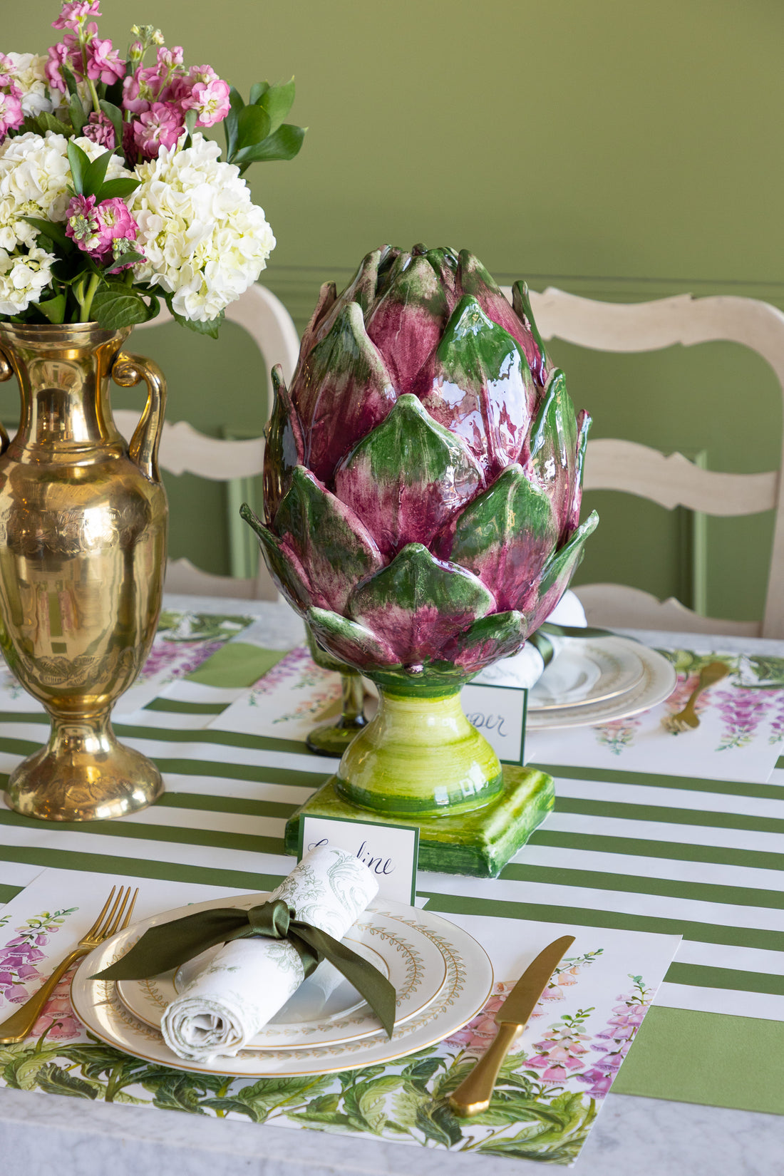 A decorative ceramic artichoke centerpiece sits on a styled dining table with green and white striped tablecloths, floral placemats, gold flatware, and a gold vase of flowers in the background.