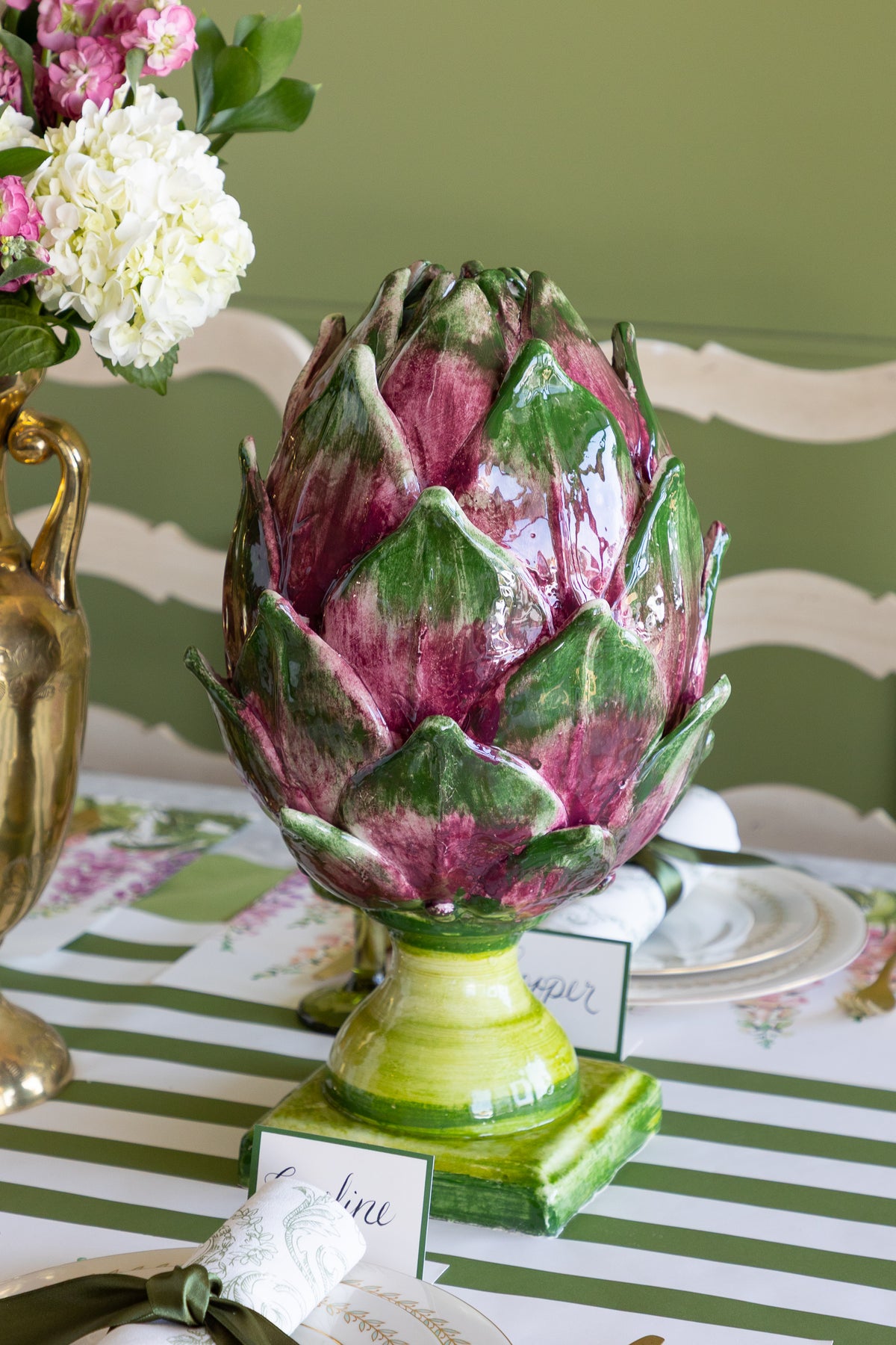A glossy ceramic artichoke centerpiece with green and deep purple-pink petals on a striped green and white tablecloth, with flowers and plates in the background.