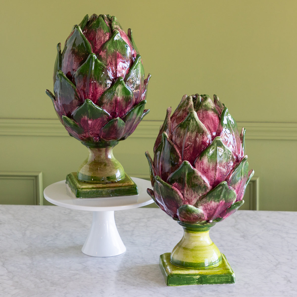 Two ceramic artichoke sculptures with green and magenta glaze, one displayed on a white pedestal stand and the other on a marble surface, set against a green wall.