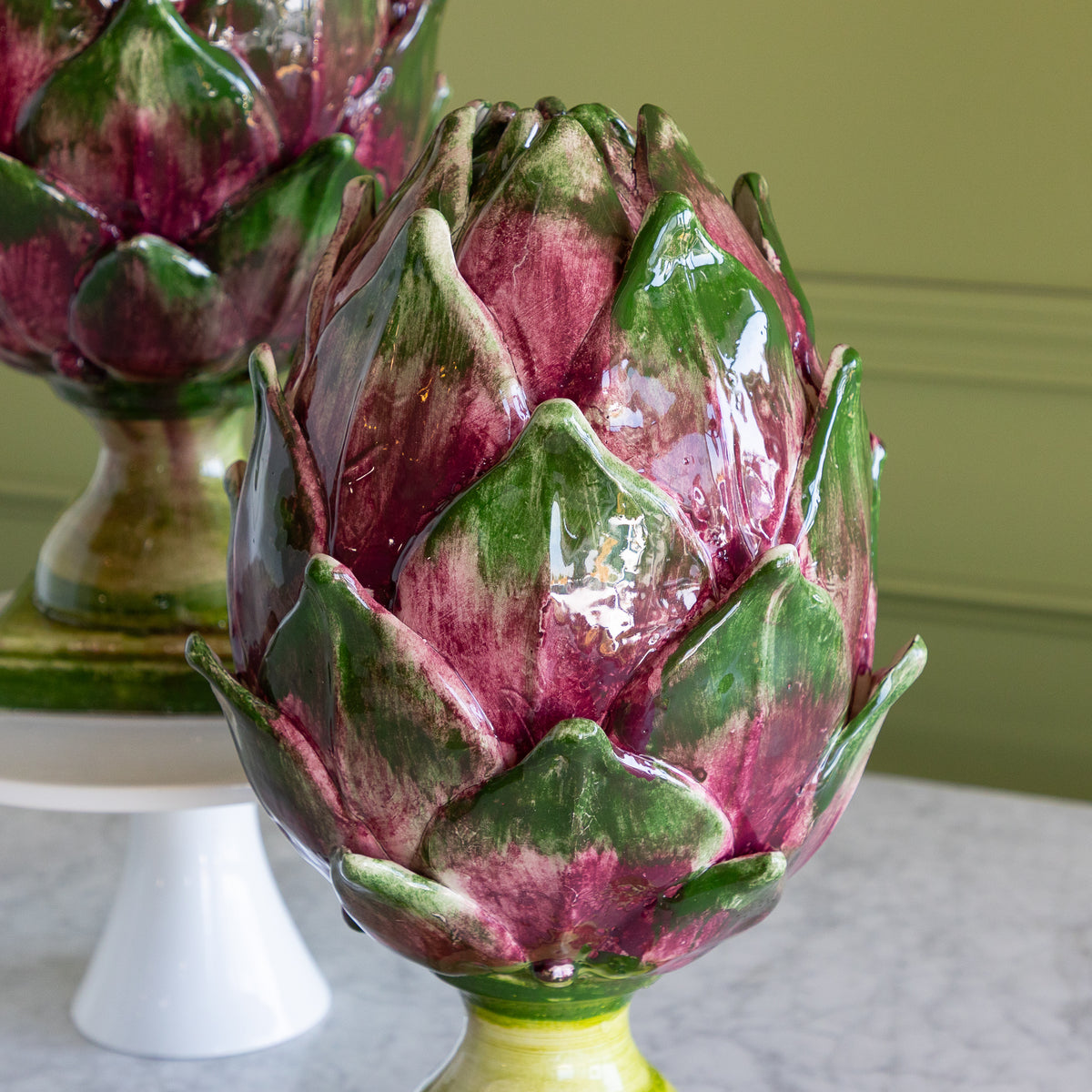 A glossy ceramic artichoke sculpture in shades of green and deep magenta, displayed on a white pedestal against an olive green wall.