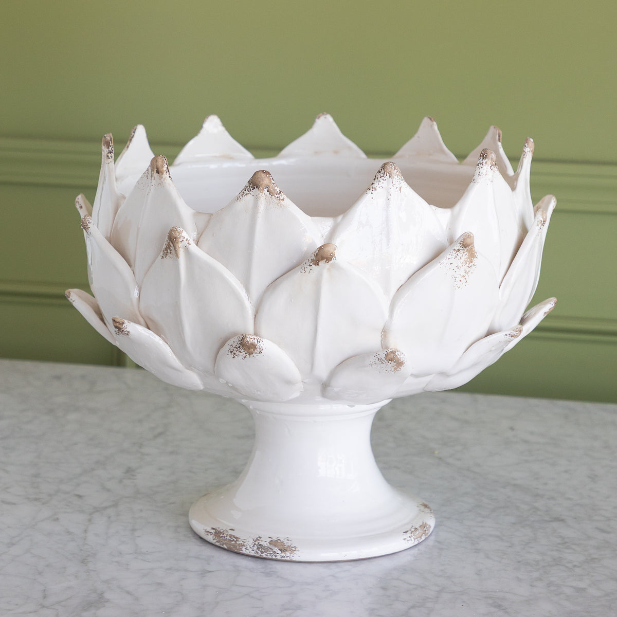 A white ceramic pedestal bowl  with an artichoke design, shown on a marble surface against a sage green paneled wall.