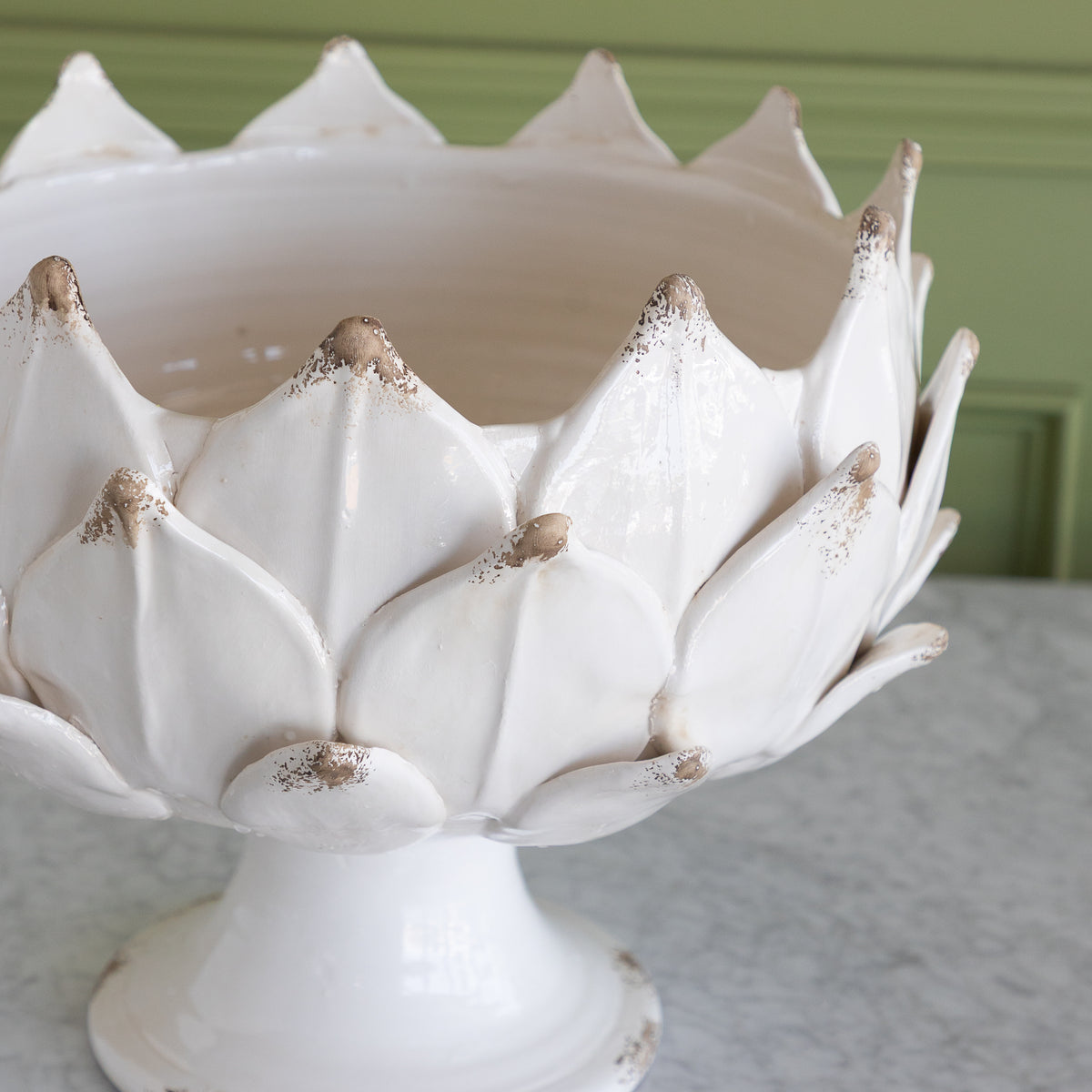 A white ceramic pedestal bowl designed like an artichoke, featuring distressed brown speckles on the petal tips, set on a light marble surface against a green paneled wall.