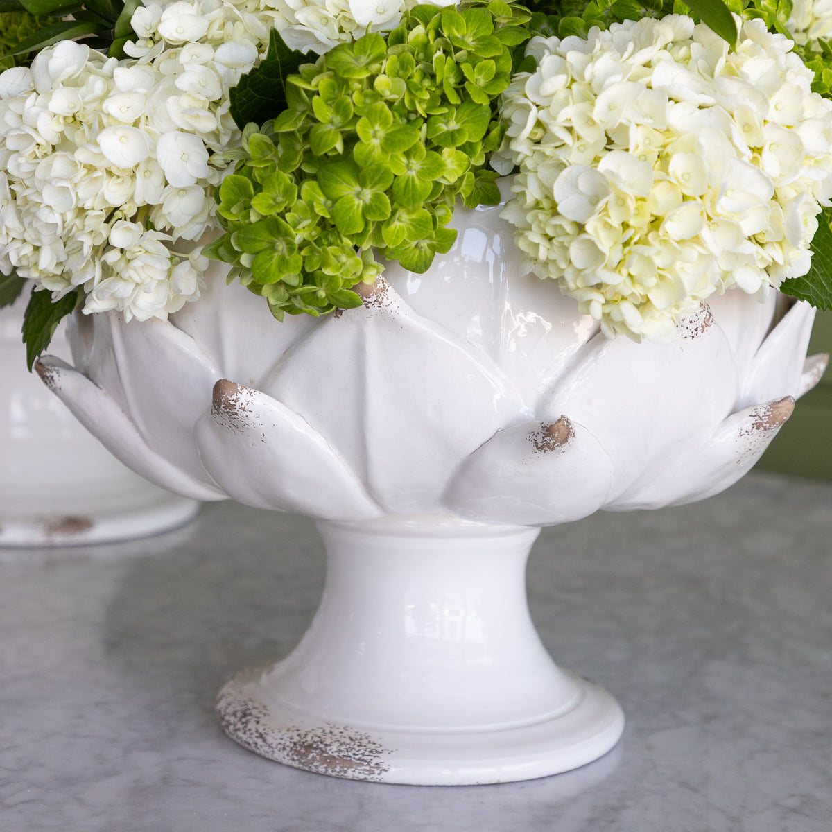 A white ceramic pedestal bowl with an artichoke design, filled with white and green hydrangea flowers, set on a light gray marble surface.
