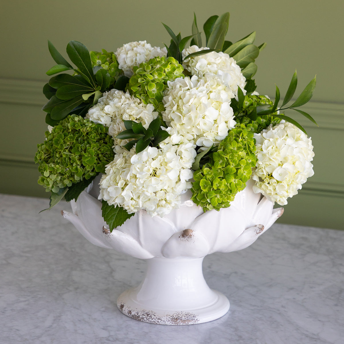 A white ceramic pedestal bowl filled with a floral arrangement of white and green hydrangeas, set on a light marble surface against a muted green wall.