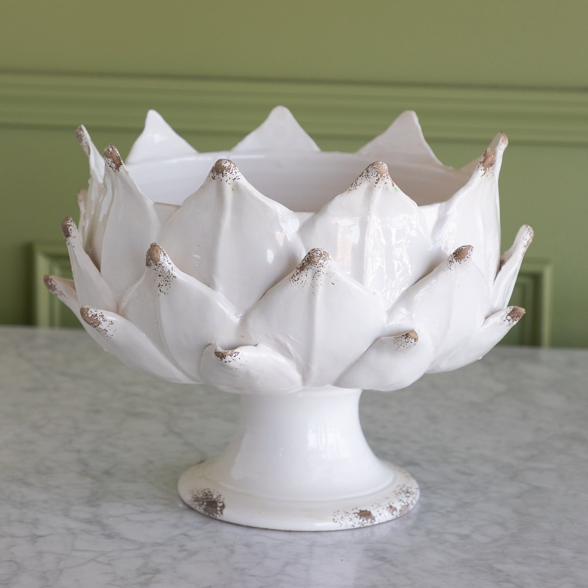 A white ceramic artichoke-shaped pedestal bowl with distressed brown speckles, set on a marble surface against a green paneled wall.