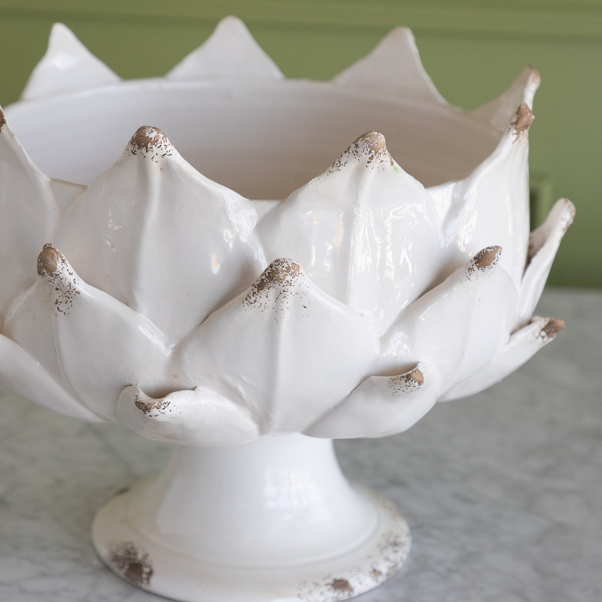 A white ceramic artichoke pedestal bowl with distressed brown speckles on the petal tips, set on a marble surface against a green wall.