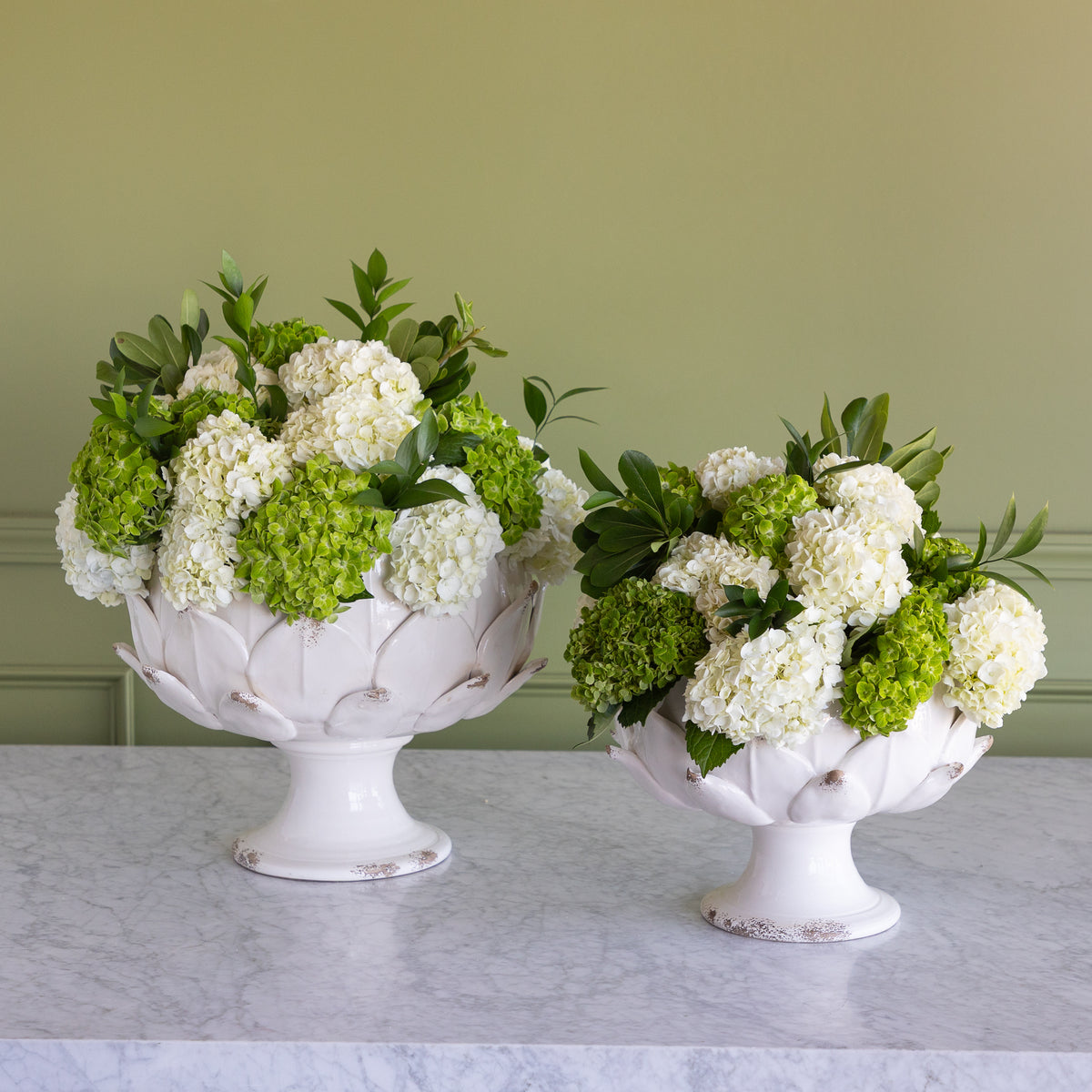 Two white pedestal flower bowls filled with white and green hydrangea arrangements, displayed on a light marble surface against a sage green paneled wall.
