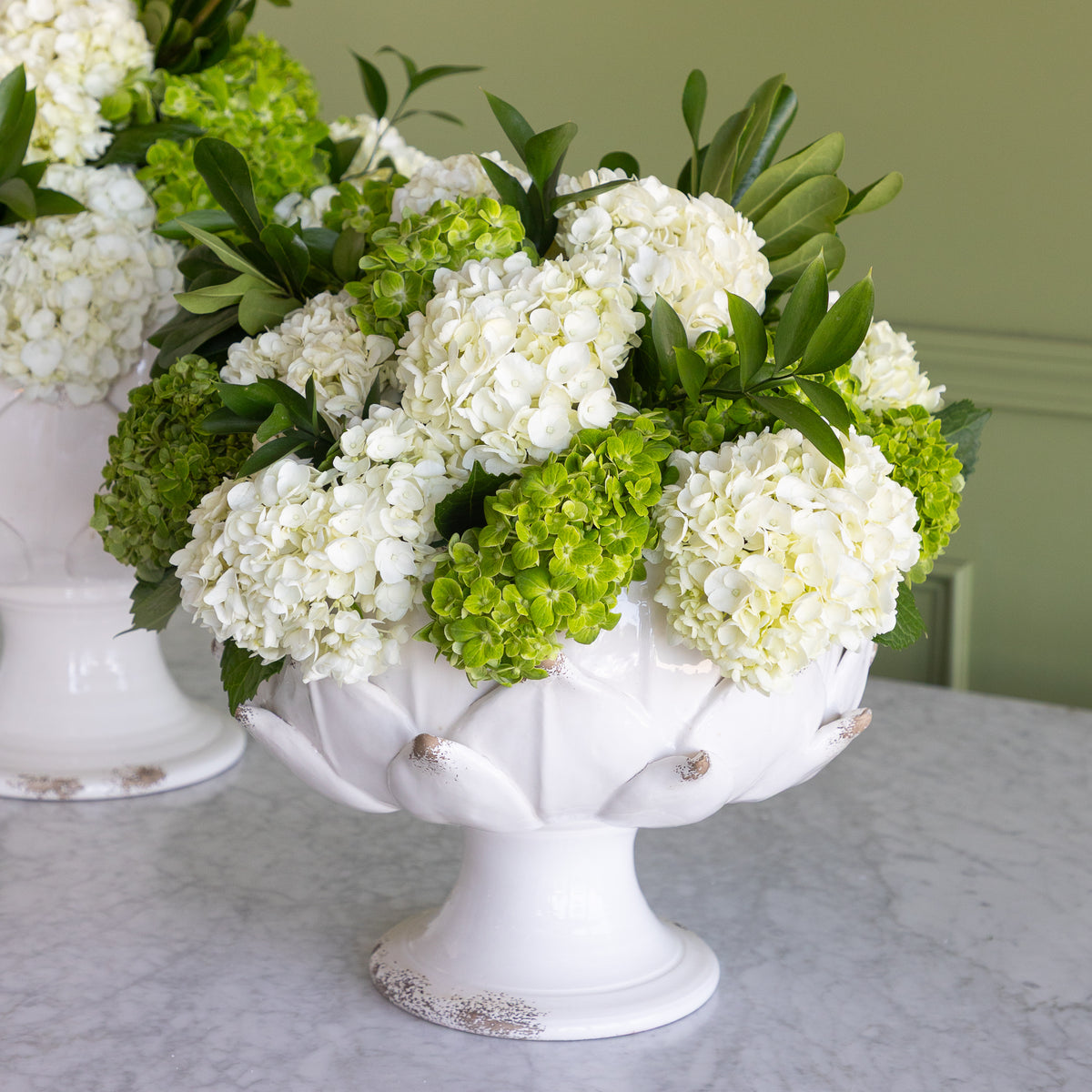 A white pedestal bowl filled with white and green hydrangea flowers, displayed on a light marble tabletop against a soft green wall.