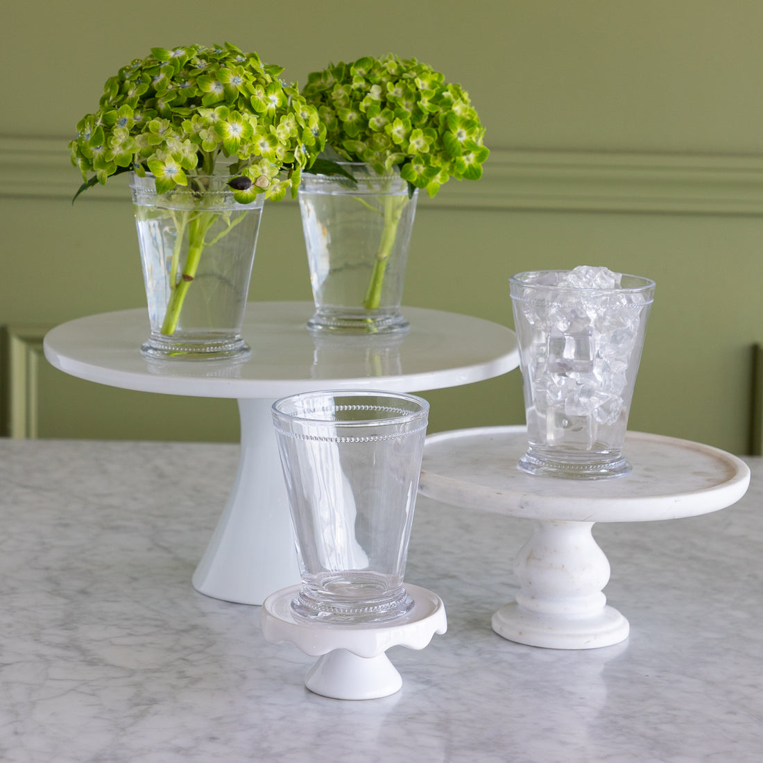 Three white pedestal cake stands arranged on a marble countertop, with two clear drinking glasses on the stands and two green hydrangea arrangements in tall clear glasses in the background.