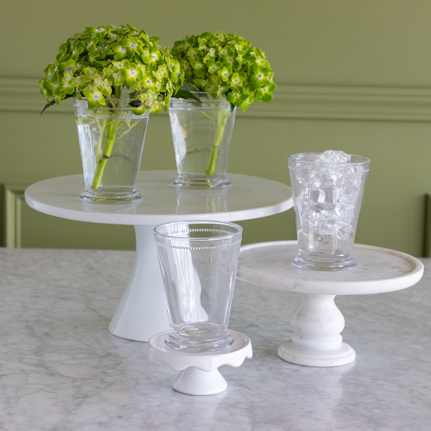 Three white pedestal cake stands arranged on a marble countertop, with two clear drinking glasses on the stands and two green hydrangea arrangements in tall clear glasses in the background.
